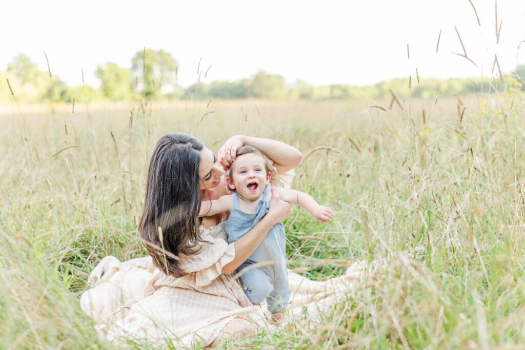 mother brushes sons hair back as they sit in field of tall grass during one year old family photo session with Sara Sniderman Photography at Heard Farm in Wayland Massachusetts