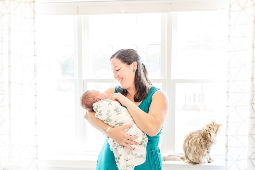 mom stand by window with newborn and cat during in home newborn session with Sara Sniderman Photography in Medway Massachusetts