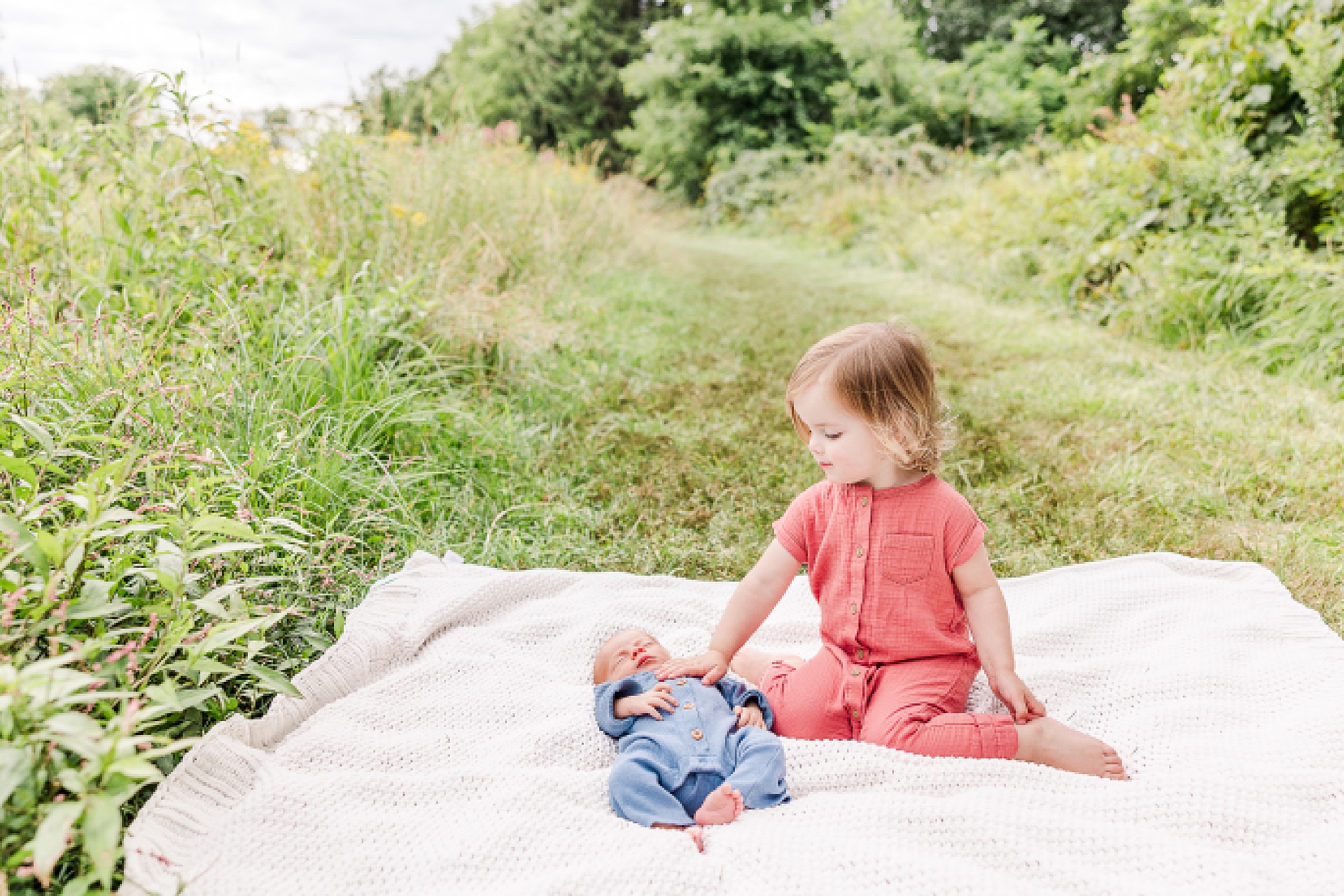 big sister sits with newborn on a blanket in a field during outdoor newborn photo session with Sara Sniderman Photography in Natick Massachusetts