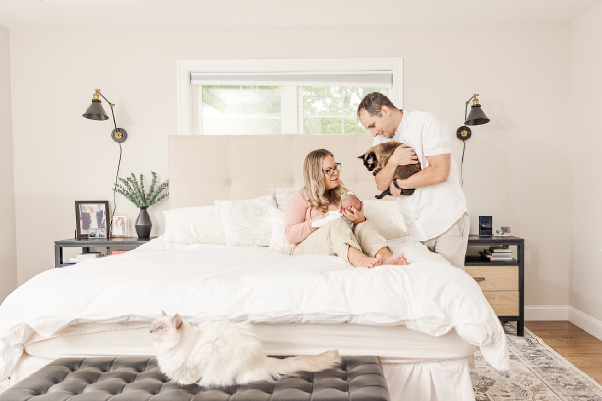 mom sits on bed with newborn an dad stands holding cat during in home newborn photo session with Sara Sniderman Photography in Natick Massachusetts