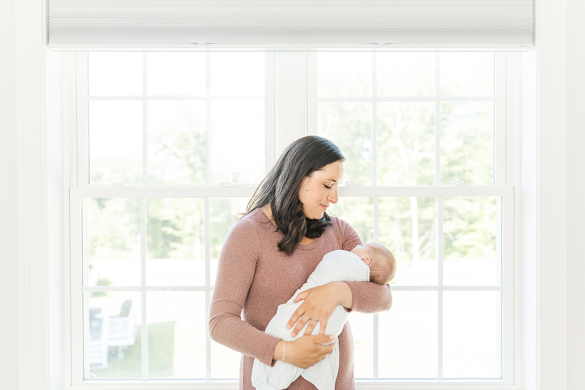 Mom holds baby in front of window during Newborn photos with Sara Sniderman Photography in Natick Massachusetts