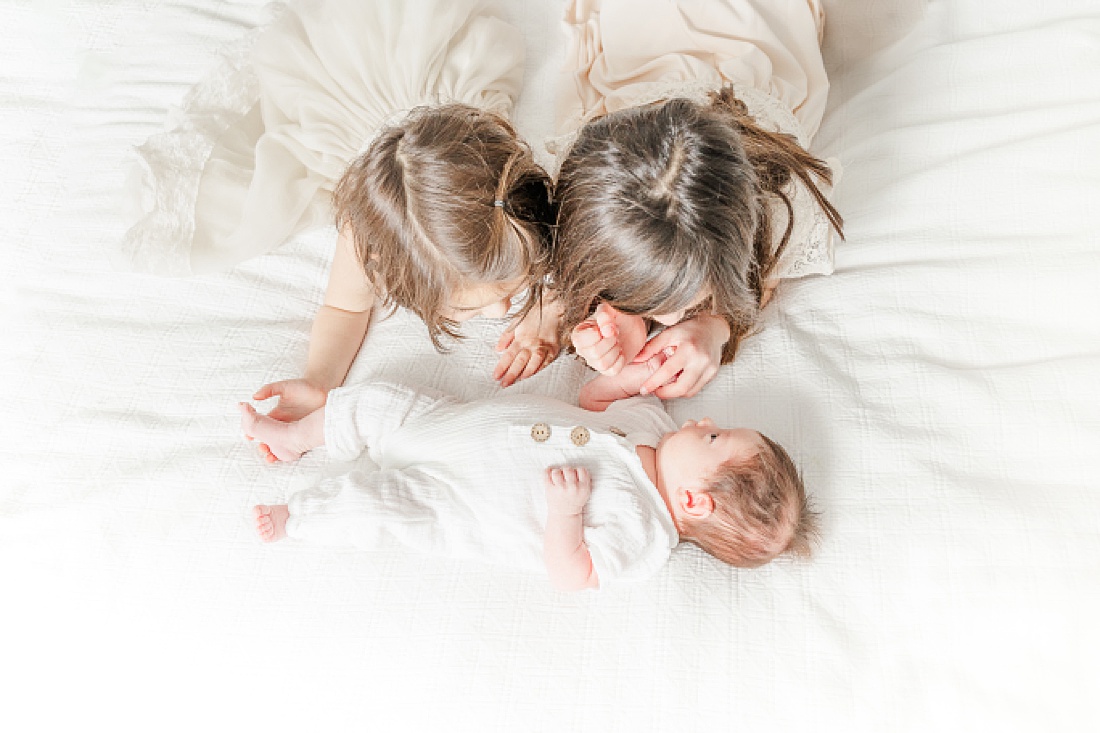 sisters lay on bed with baby during in-home newborn photo session with Sara Sniderman Photography in Needham Massachusetts