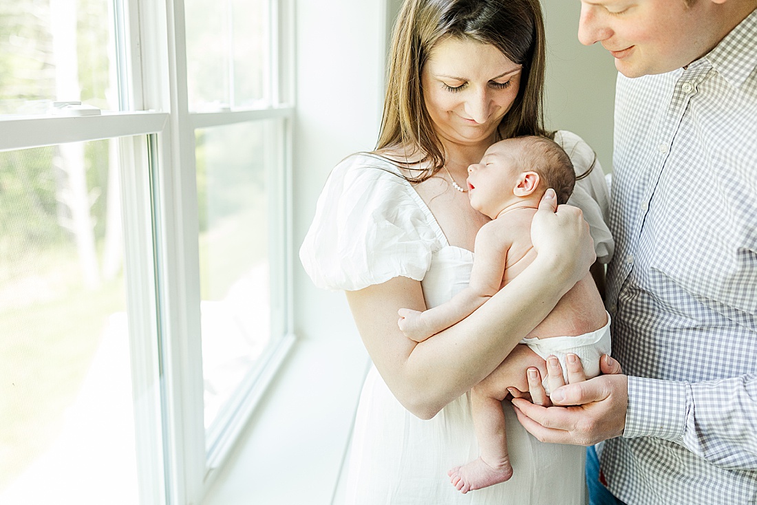 mother holds newborn by window during in-home photo session in Millis Massachusetts with Sara Sniderman Photography