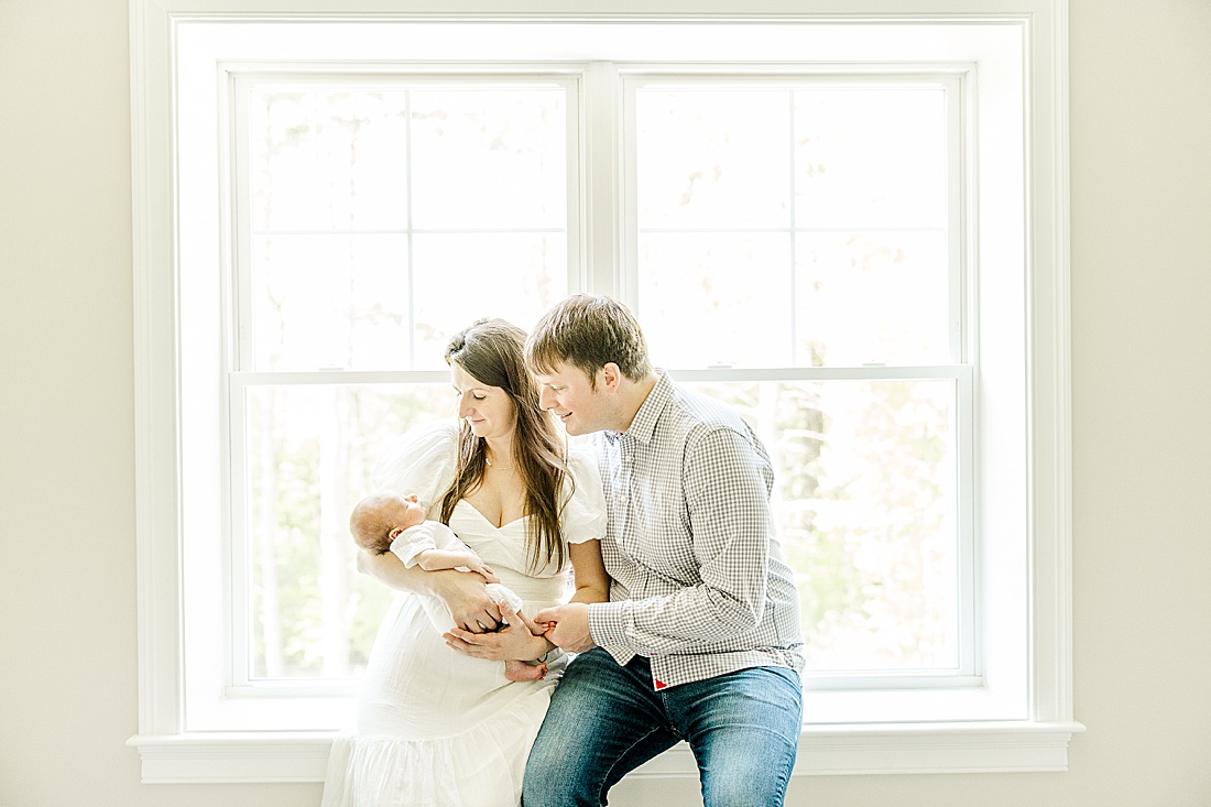 parents sit by window with baby during in-home photo session in Millis Massachusetts with Sara Sniderman Photography