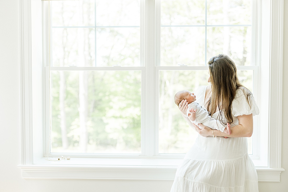 mom sits by window with baby during in-home photo session in Millis Massachusetts with Sara Sniderman Photography