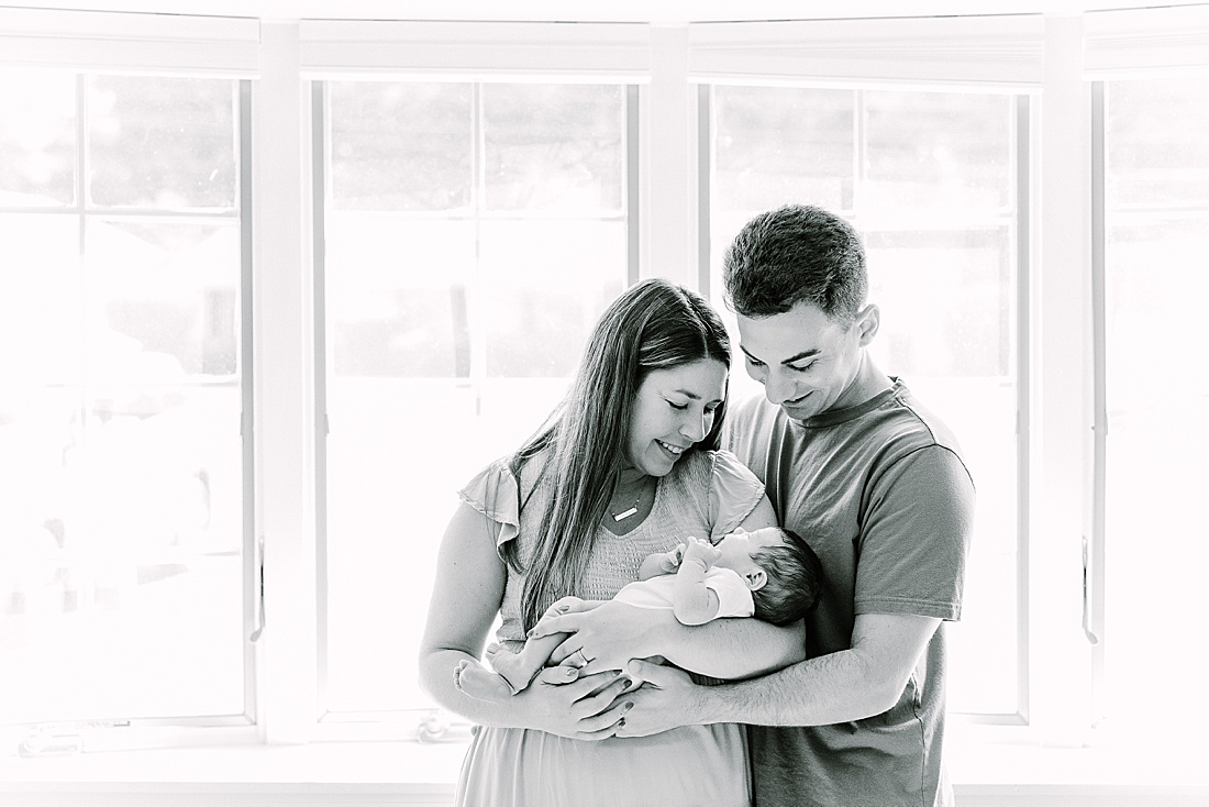 Image of professional newborn photos taken by Sara Sniderman Photography, a family, newborn and maternity photographer serving the Metro West Boston area include: Natick, Needham, Newton, Wellesley, Wayland, Weston, Sherborn, Sudbury, Southborough, Medfield, Medway, Holliston, Framingham. Image shows black and white photo of parents holding baby by a window.