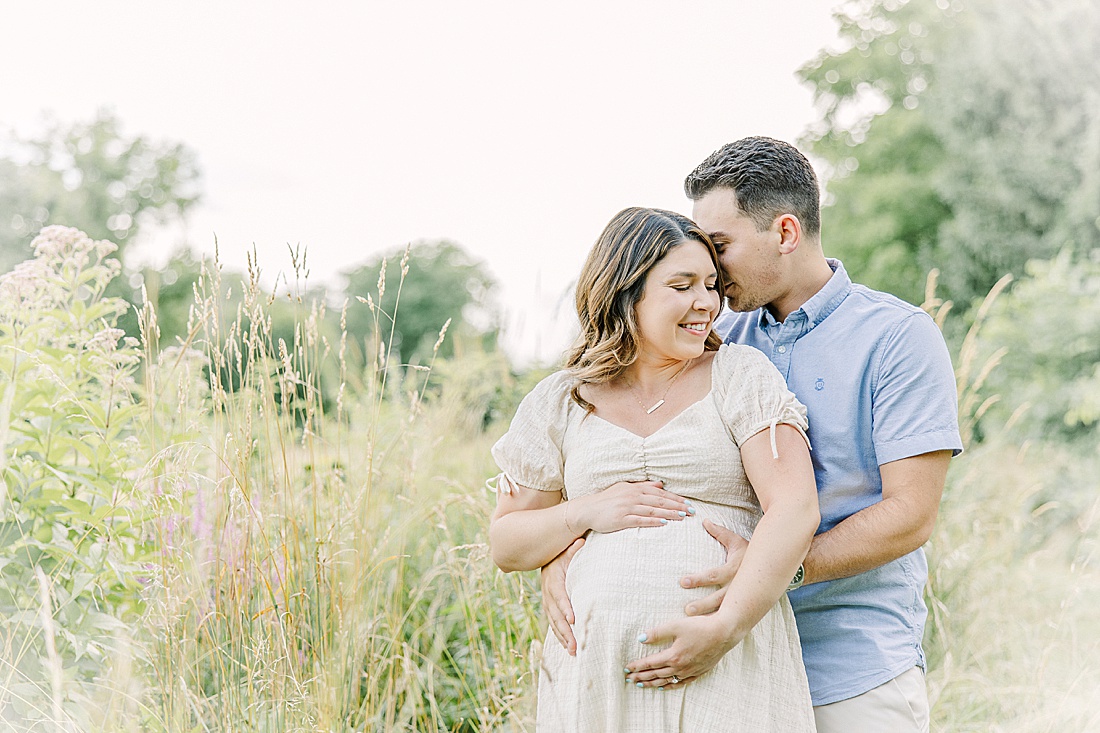 Image of professional maternity photos taken by Sara Sniderman Photography, a family, newborn and maternity photographer serving the Metro West Boston area include: Natick, Needham, Newton, Wellesley, Wayland, Weston, Sherborn, Sudbury, Southborough, Medfield, Medway, Holliston, Framingham. Image shows husband and wife holding belly in field of tall grass