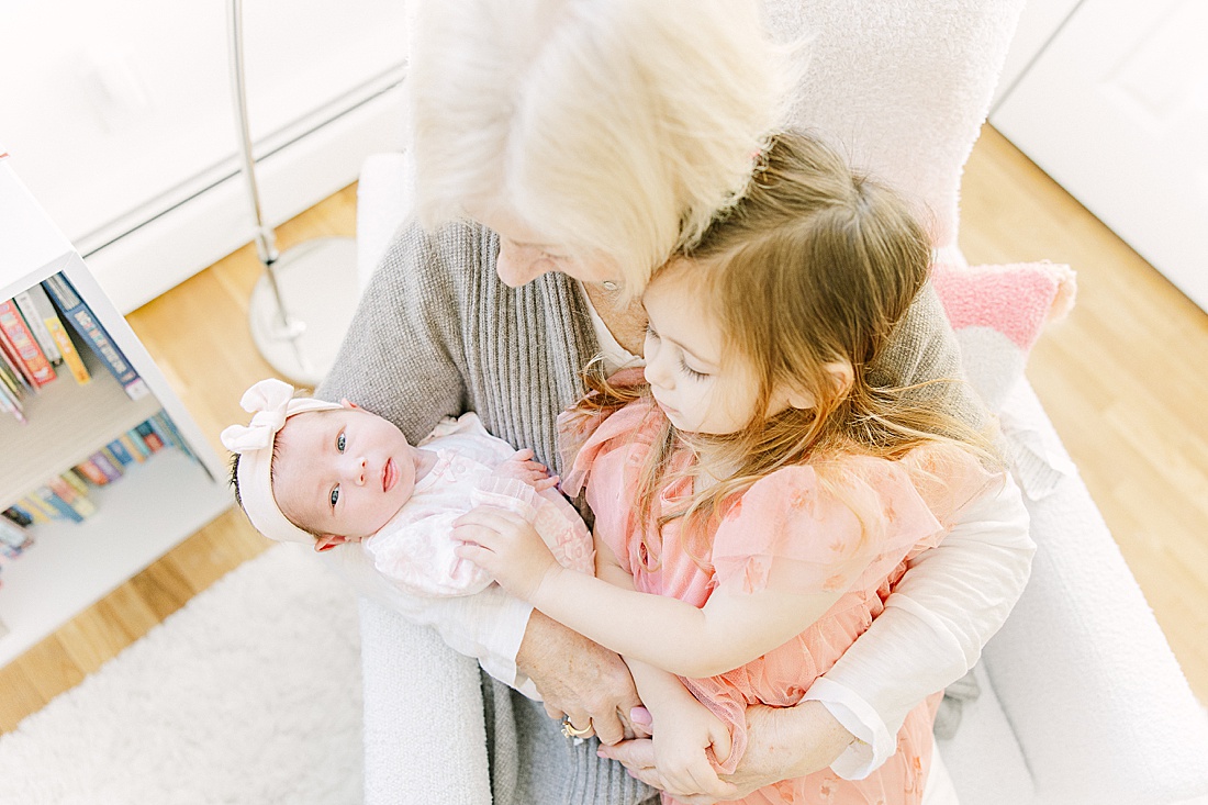 grandmother sits in chair with toddler and baby during in-home newborn photo session with Sara Sniderman Photography in Needham Massachusetts