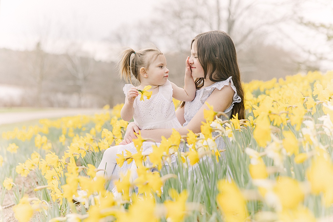 sisters sit in daffodil field during first birthday photo session with Sara Sniderman Photography in Wellesley Massachusetts