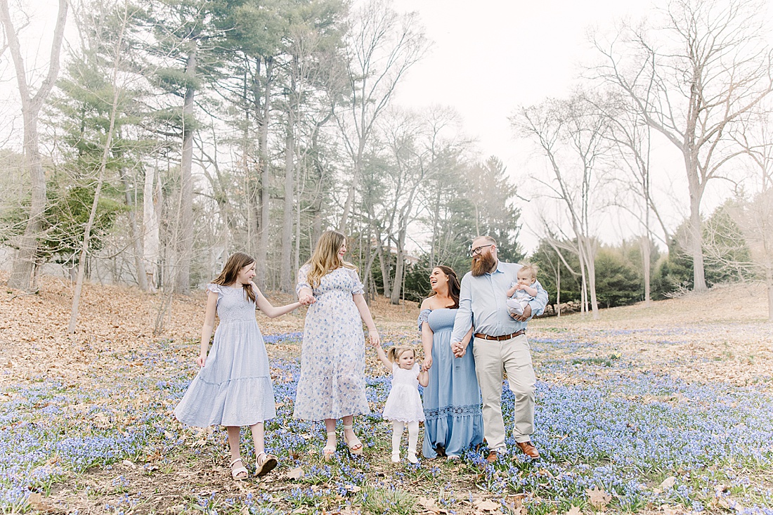 family walks through field of blue flowers during first birthday photo session with Sara Sniderman Photography in Wellesley Massachusetts