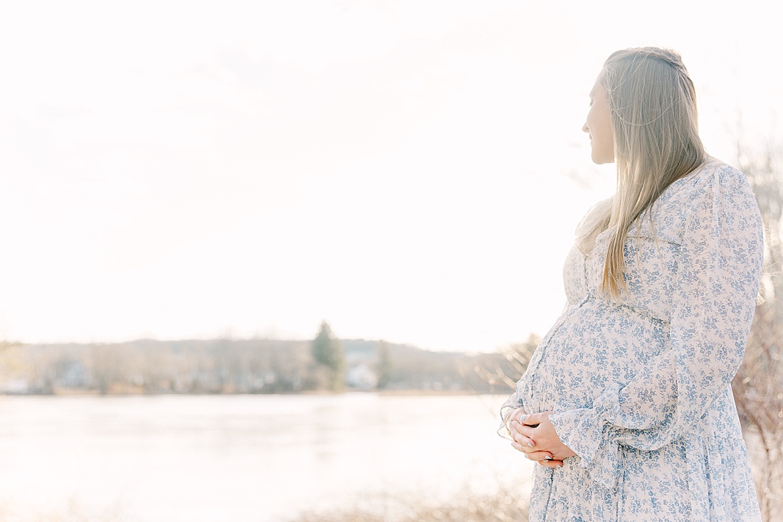pregnant woman holds bump and looks out over the Ashland Reservoir during maternity photos session with Sara Sniderman Photography in Ashland Massachusetts