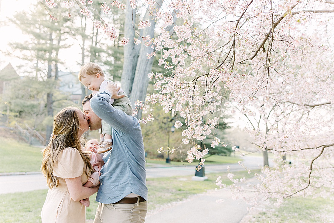 parents kiss while holding kids uring family photo session with Sara Sniderman Photography at Wellesley College.