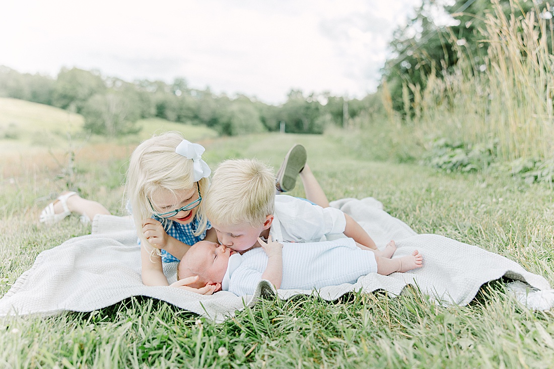 older siblings kiss baby laying on blanket during outdoor photo session with Sara Sniderman Photography at Breakneck Hill Southborough Massachusetts