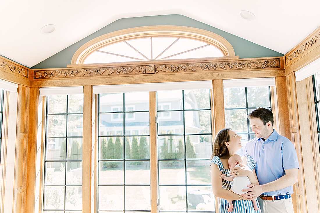 Parents hold baby in front of wall of windows during in-home newborn photo session with Sara Sniderman Photography in Wellesley Massachusetts. 