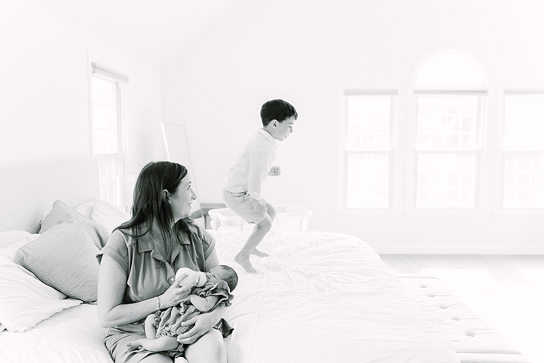 black and white photo of mother feeding baby and big brother jumping on the bed during in-home newborn photo session with Sara Sniderman Photography in Sherborn Massachusetts