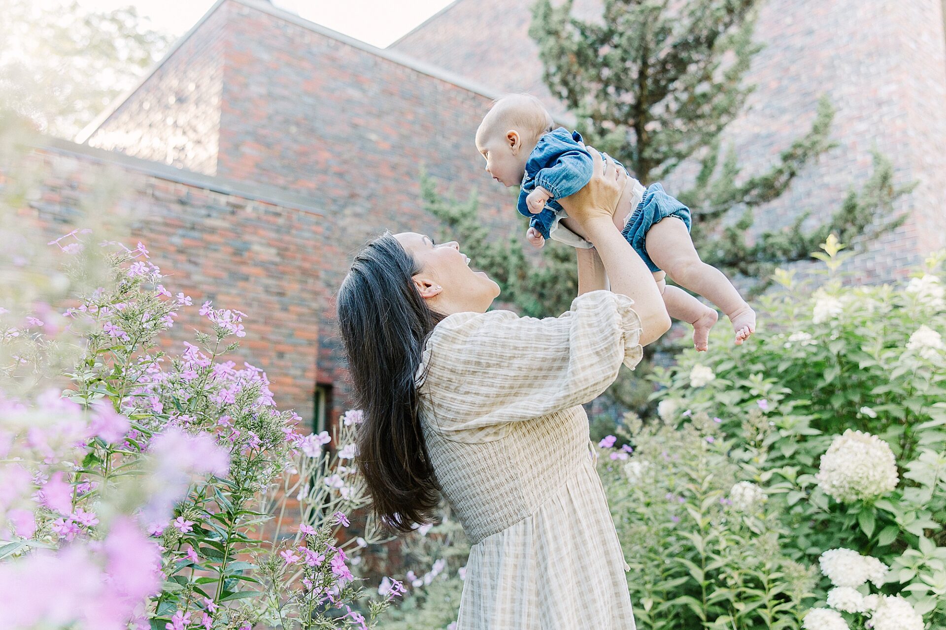 mom stands next too flower bush holding baby up high during outdoor newborn photo session with Sara Sniderman Photography at Wellesley College.faceless baby photos